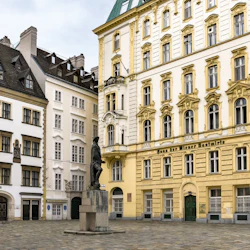 En staty står på ett kullerstensbelagt torg omgivet av utsmyckade, flervåningsbyggnader med olika arkitektoniska detaljer och stilar.