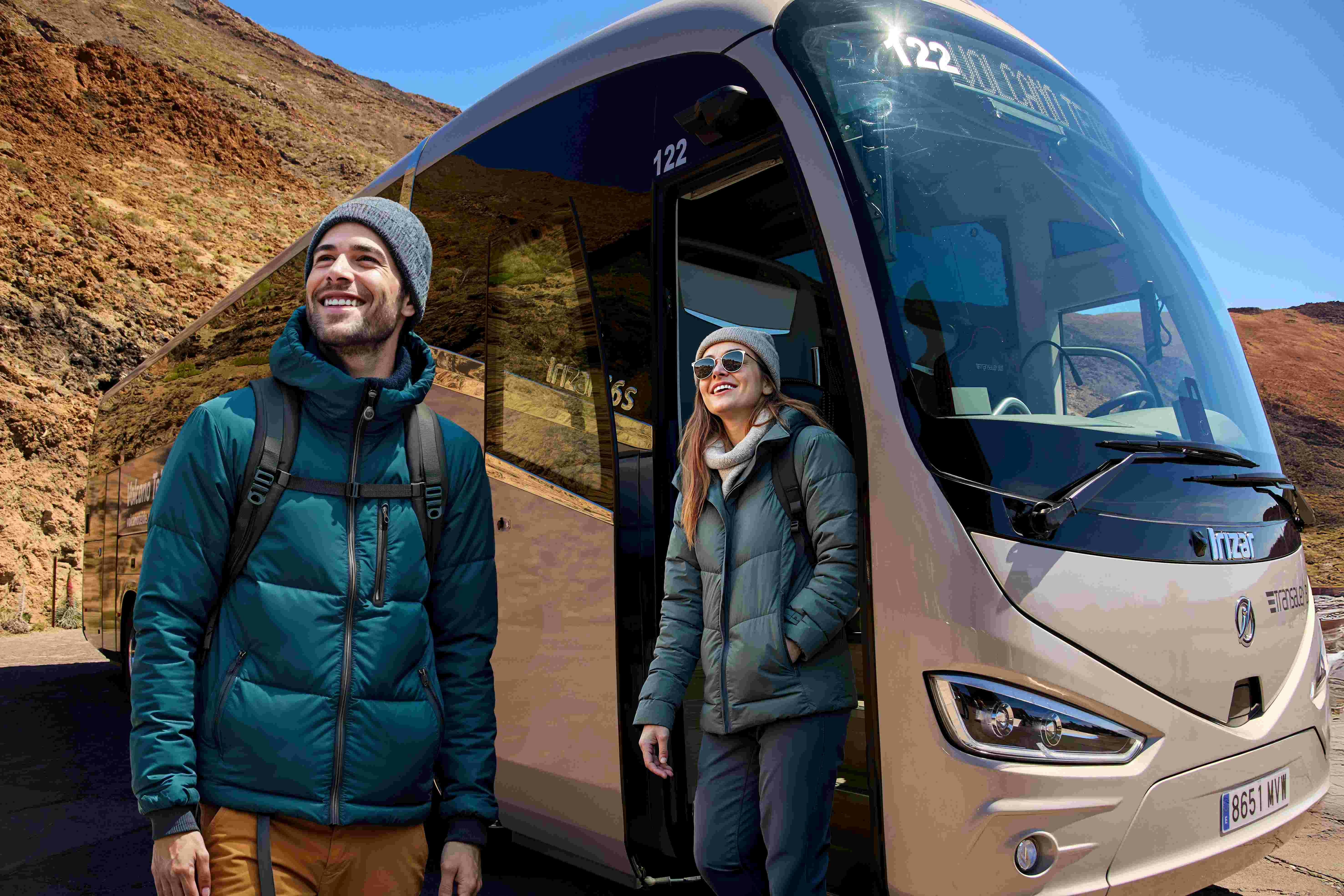 两个背着背包的人，穿着冬季夹克和帽子，站在山区的旅游巴士旁边微笑。