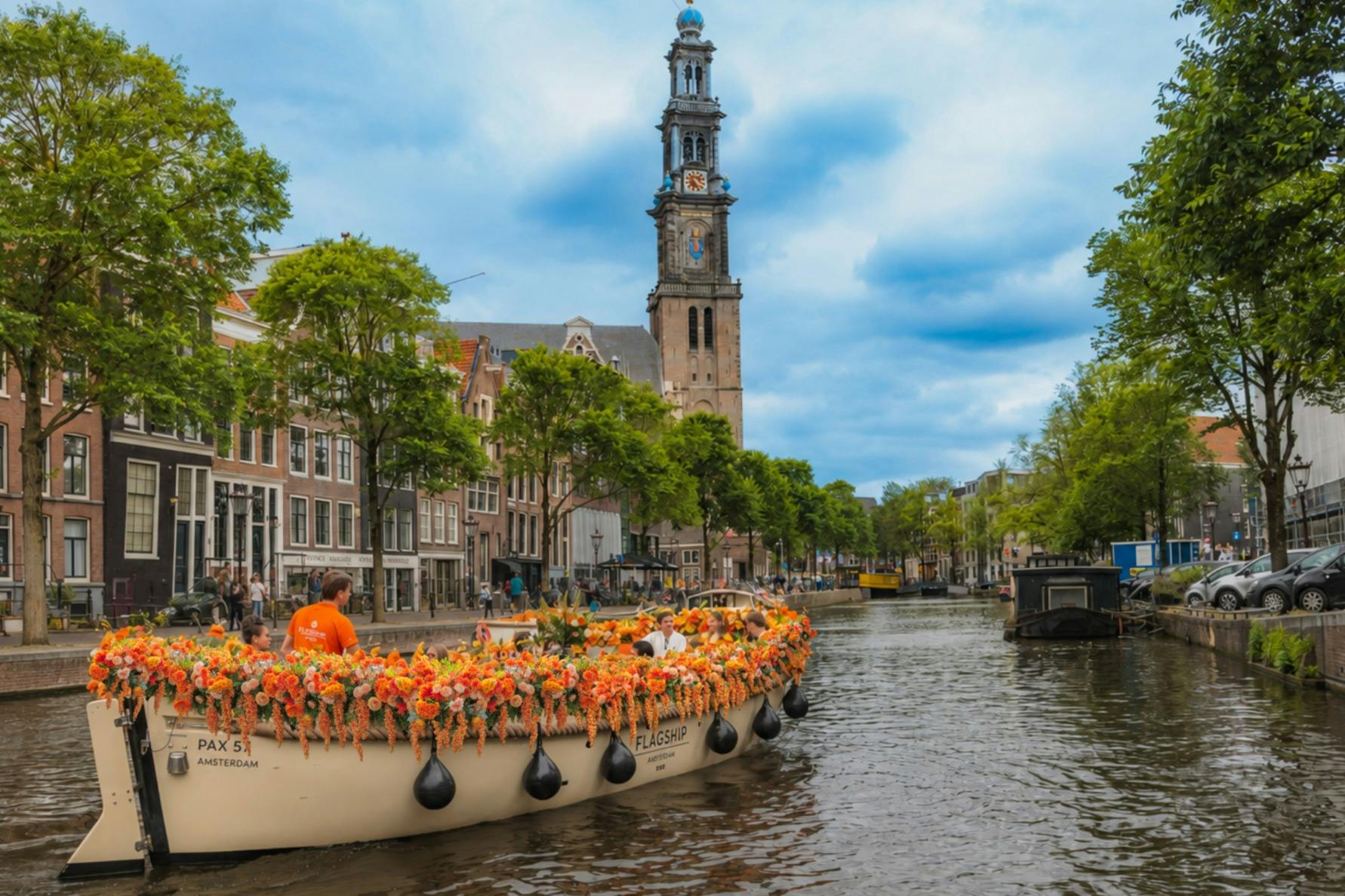 A canal in Amsterdam with a boat decorated with flowers, surrounded by historic buildings and a church tower in the background.