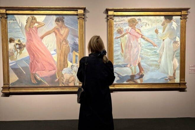 Osoba v tmavém kabátě pozorující dva zarámované obrazy představující postavy v splývavých oděvech a venkovních scénách.