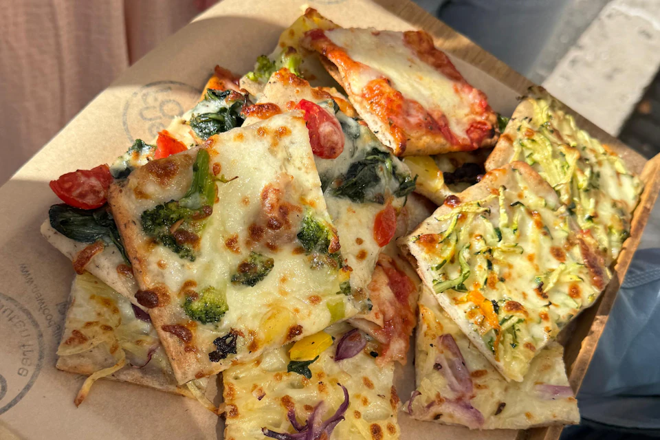Assorted slices of vegetable pizza with toppings including broccoli, tomatoes, spinach, zucchini, and cheese, arranged on brown paper.