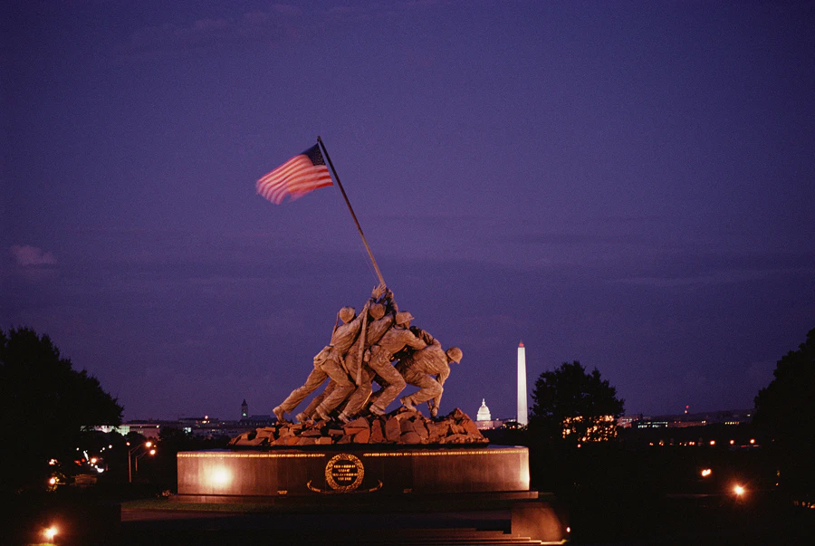 A nighttime view of the Marine Corps War Memorial depicting soldiers raising an American flag, with the Washington Monument in the background.