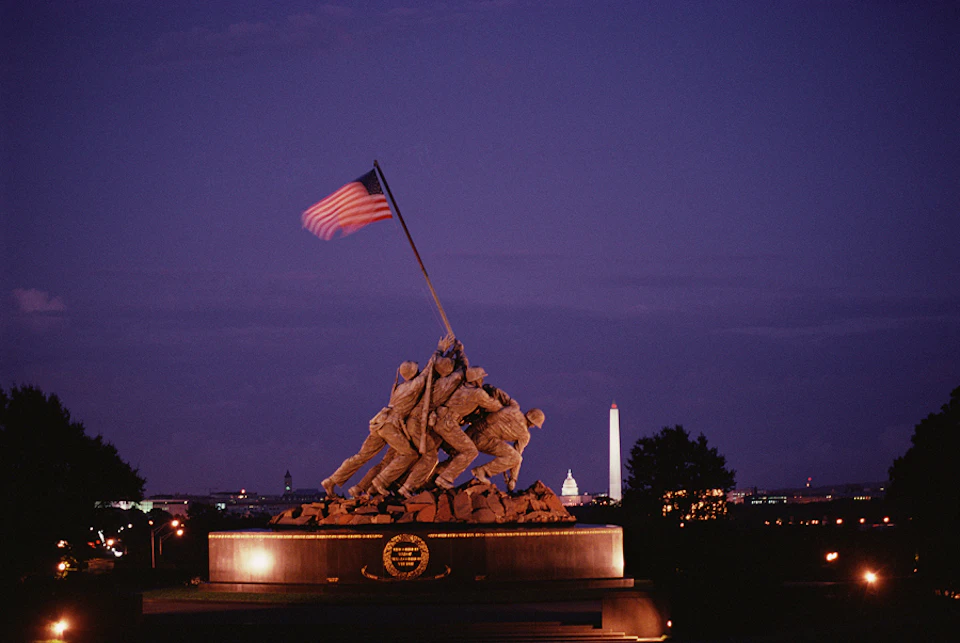 A nighttime view of the Marine Corps War Memorial depicting soldiers raising an American flag, with the Washington Monument in the background.