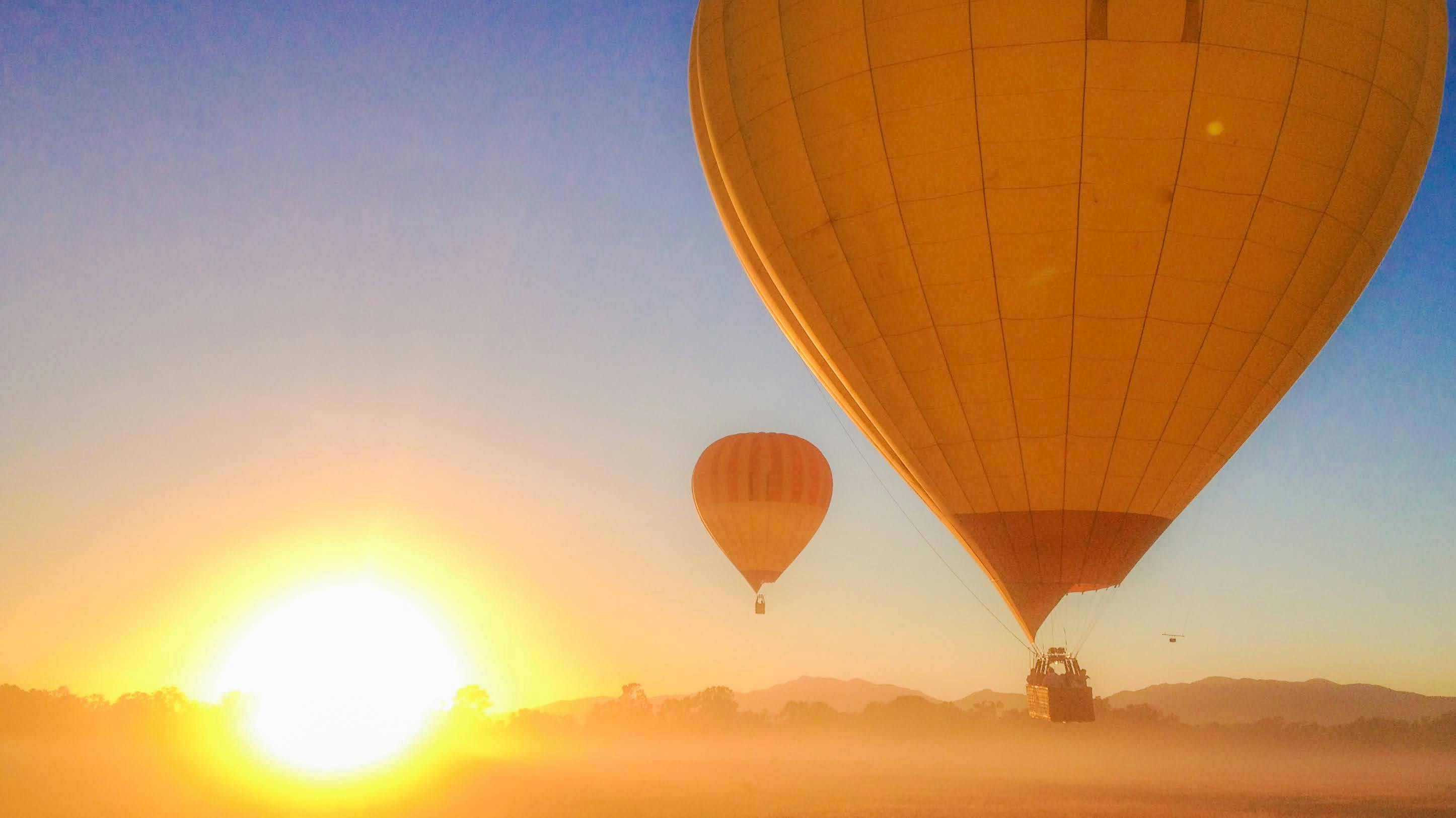 A hot air balloon floats over a sunlit town, with rays of sunlight piercing through clouds illuminating the landscape below.