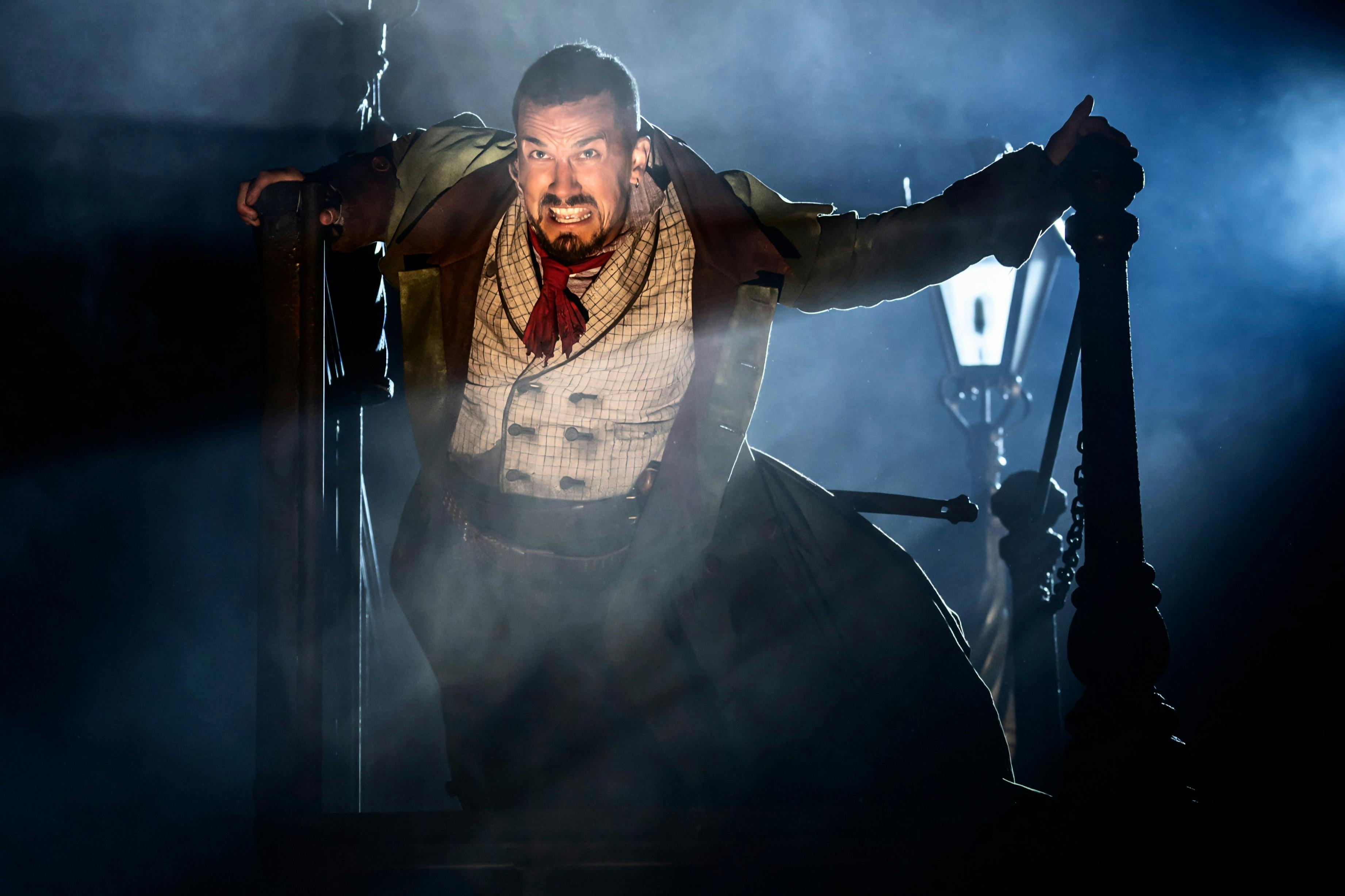 Home en disfressa d'època amb una expressió intensa, de peu en una il·luminació dramàtica, agafant barres de fusta amb una llanterna al fons.