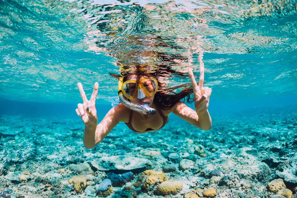 Person snorkeling underwater in clear blue water, making peace signs with both hands, with rocks and corals visible below.