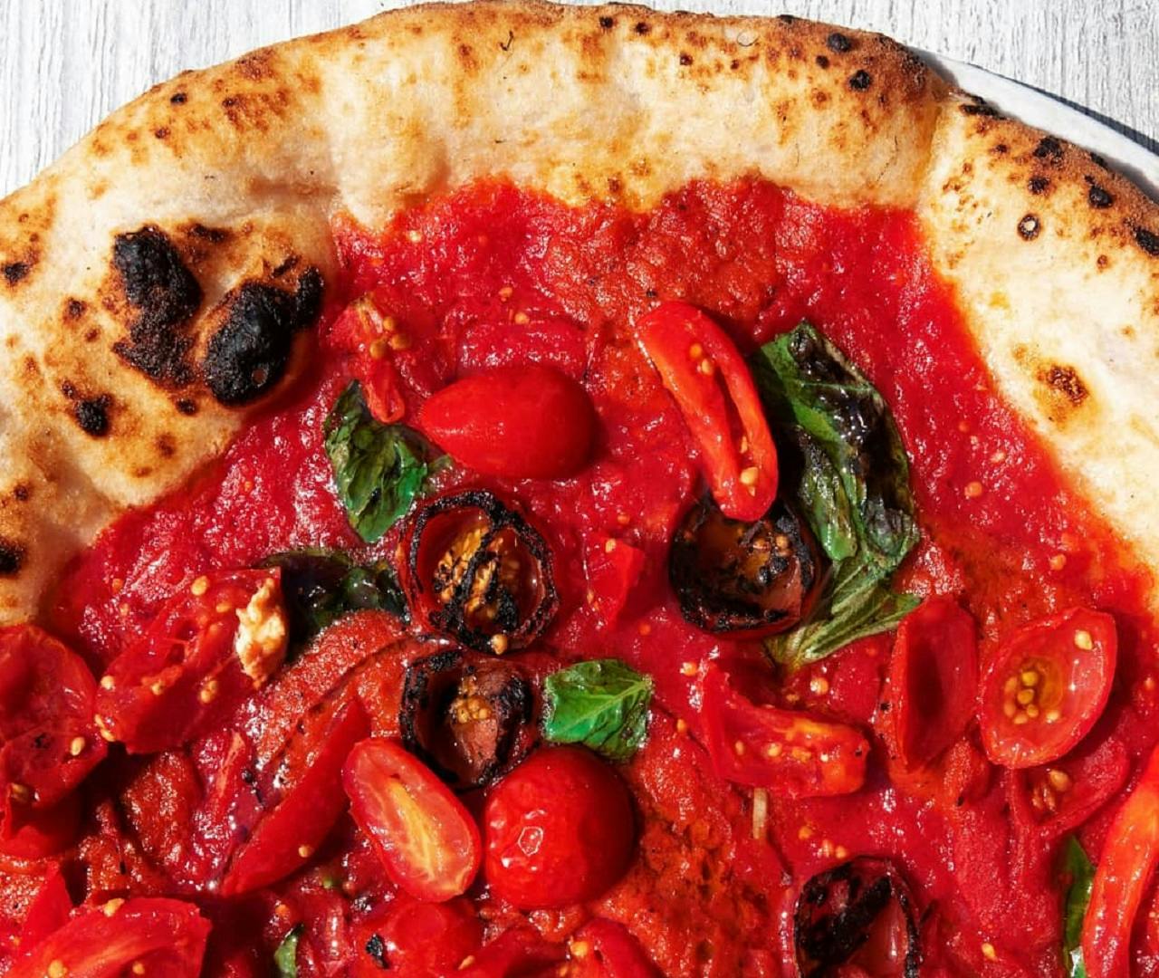 Крупный план пиццы с обугленной корочкой, увенчанной томатным соусом, помидорами черри, жареными овощами и свежим базиликом.