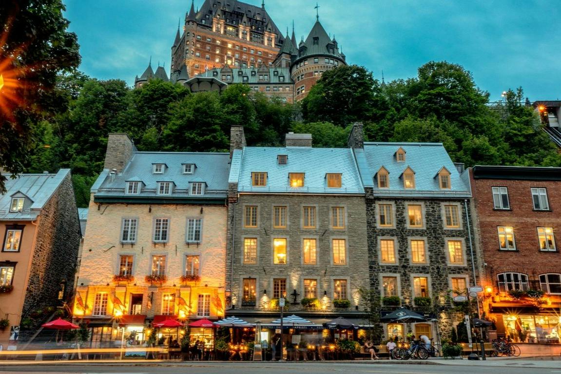 Historic European-style buildings with lit windows and a castle on a hill above, under a twilight sky.