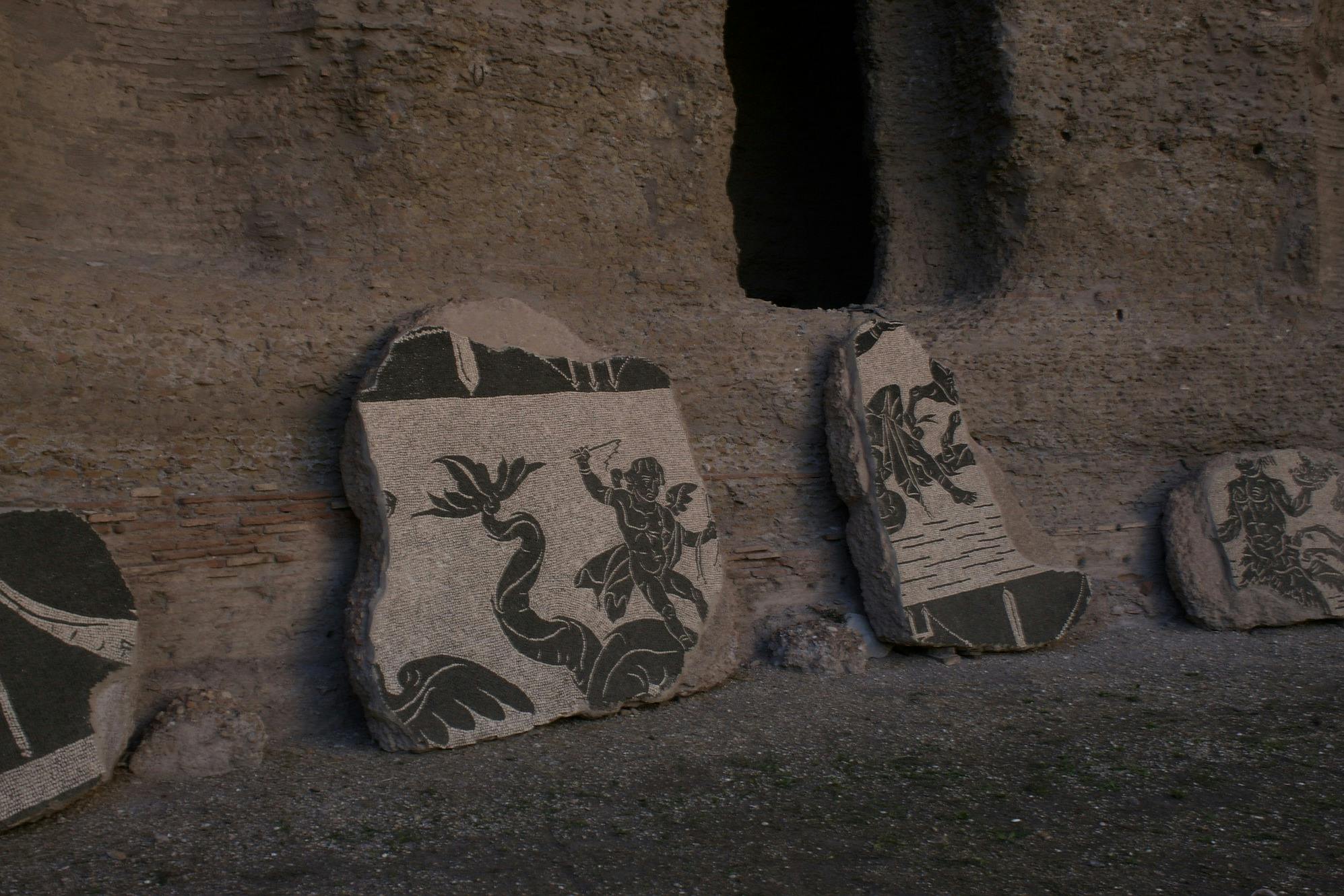 荒れた石の壁に寄りかかる黒と白のデザインの2つの古代の石のモザイク。 1つは、蛇を持つ男を描いています。