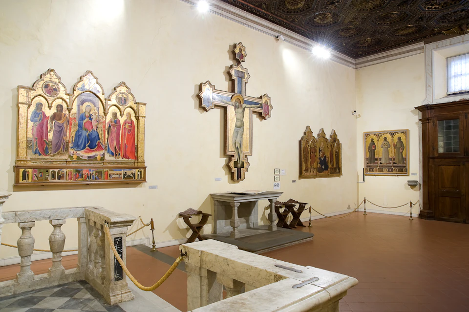 A museum room featuring religious artwork, including a large crucifix and several paintings, with a small altar and roped barriers.