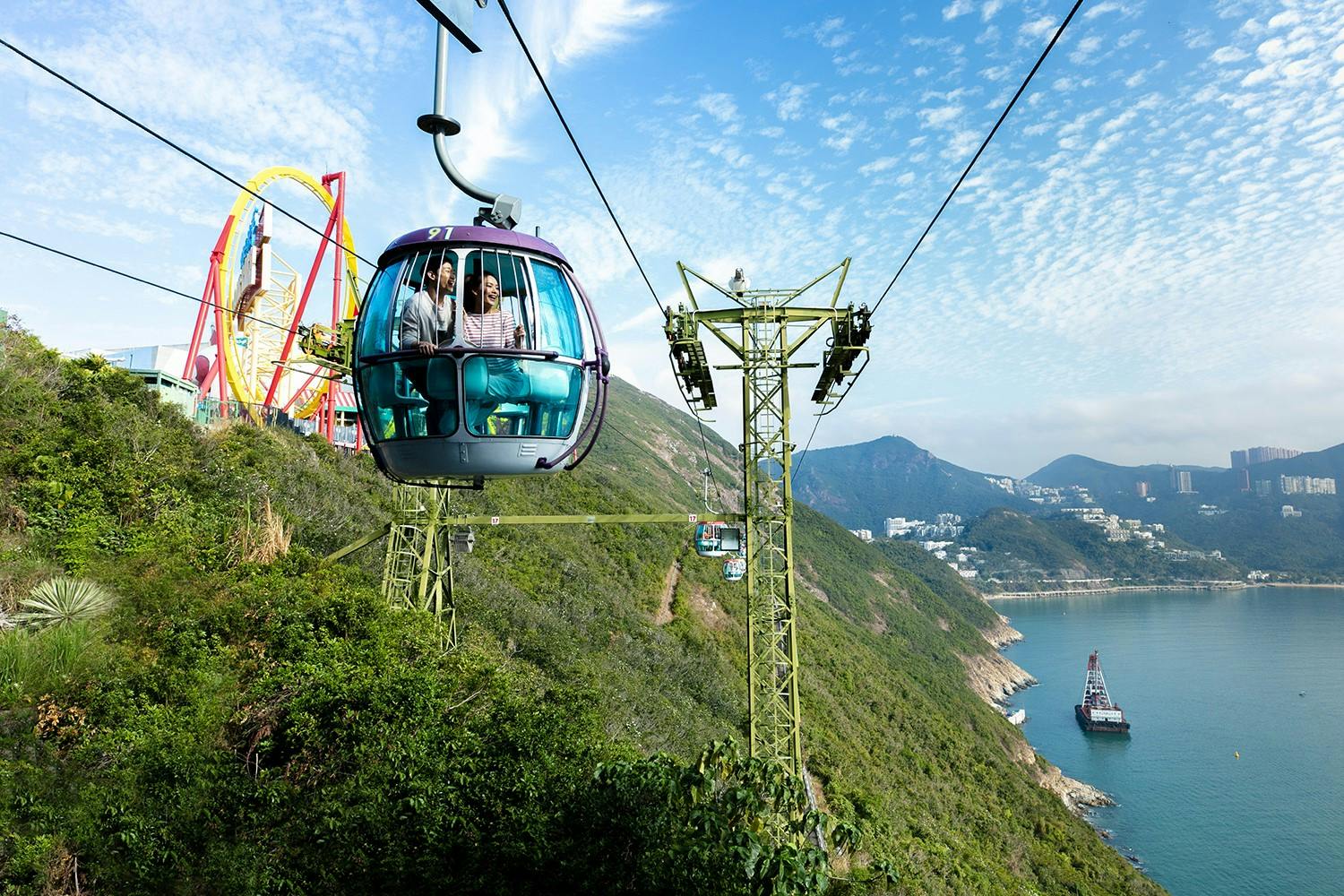 A gondola lift with passengers travels over a scenic hillside with a roller coaster and ocean in the background.