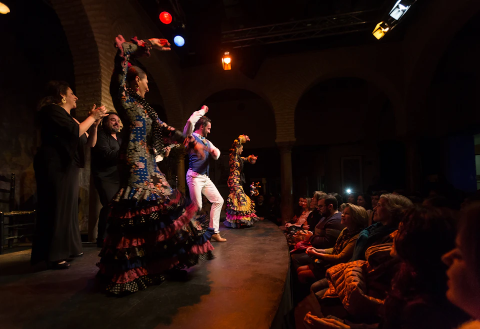 Performers in colourful dresses and suits dance flamenco on a dimly lit stage, with an audience watching them.