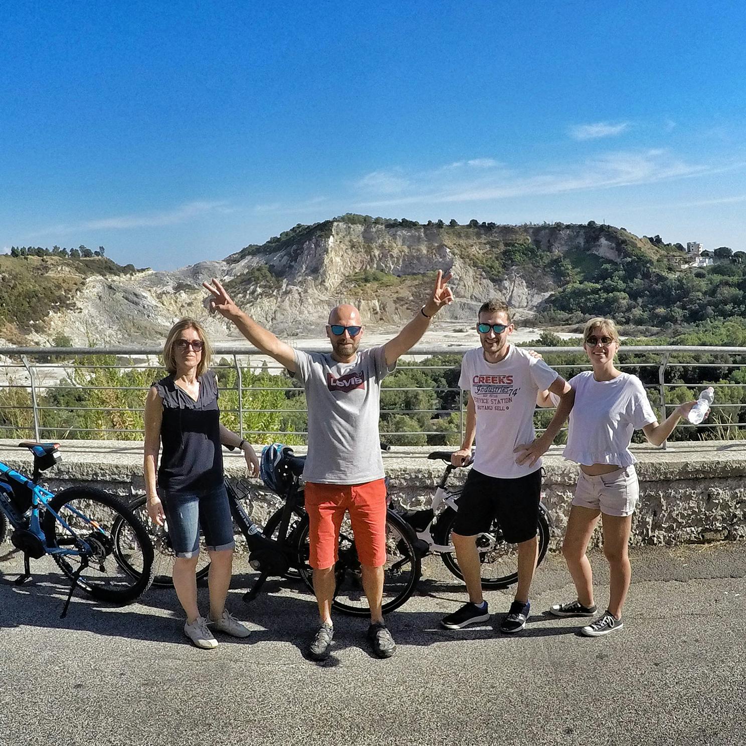 Quattro persone in piedi con le biciclette su una strada asfaltata, in posa davanti a una scenografica collina rocciosa sotto un cielo azzurro.