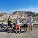 Four people stand with bicycles on a paved road, posing in front of a scenic rocky hillside under a blue sky.