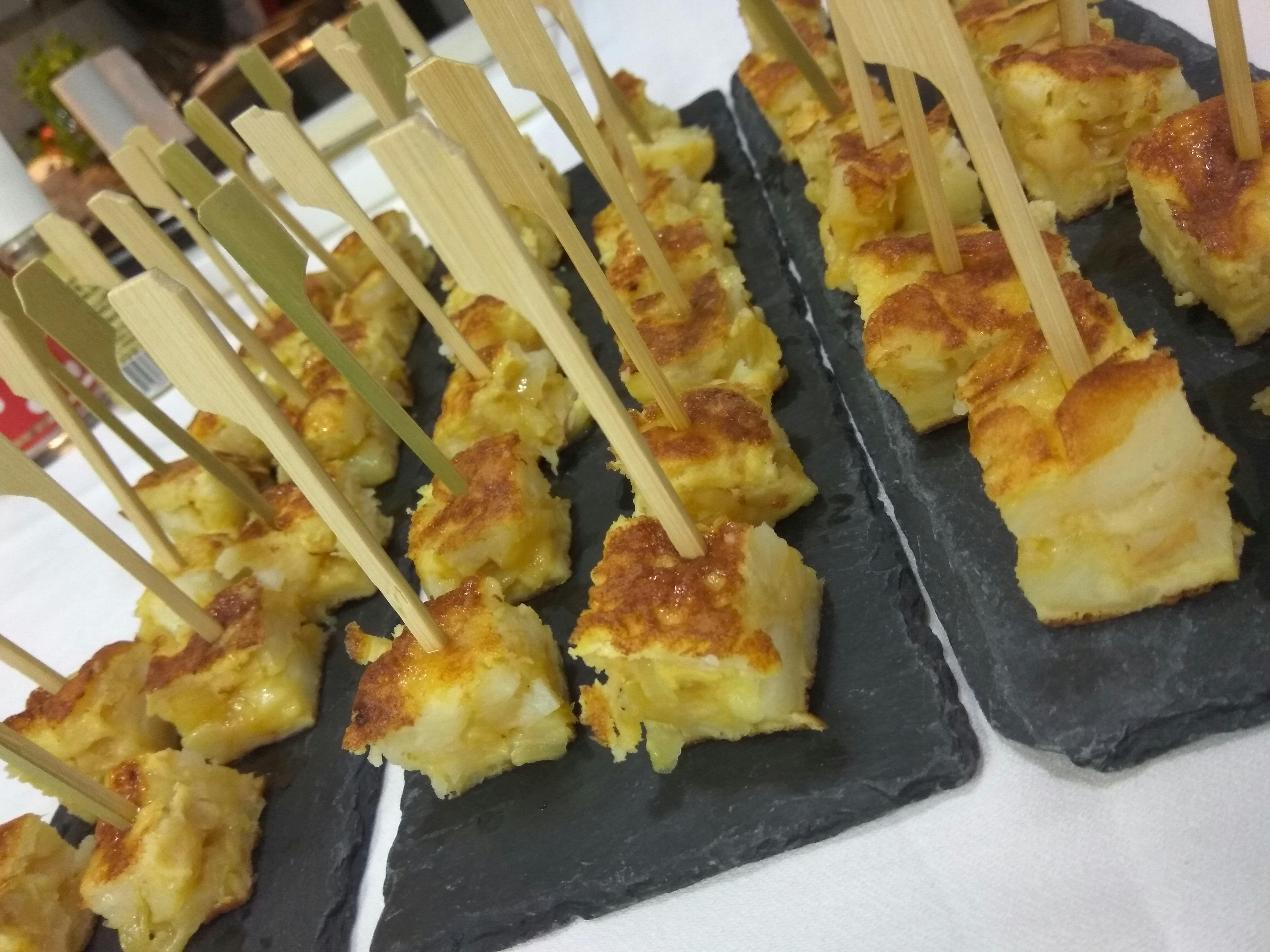 Небольшие квадратные кусочки испанской тортильи с бамбуковыми шампурами, расположенные на сланцевых тарелках.