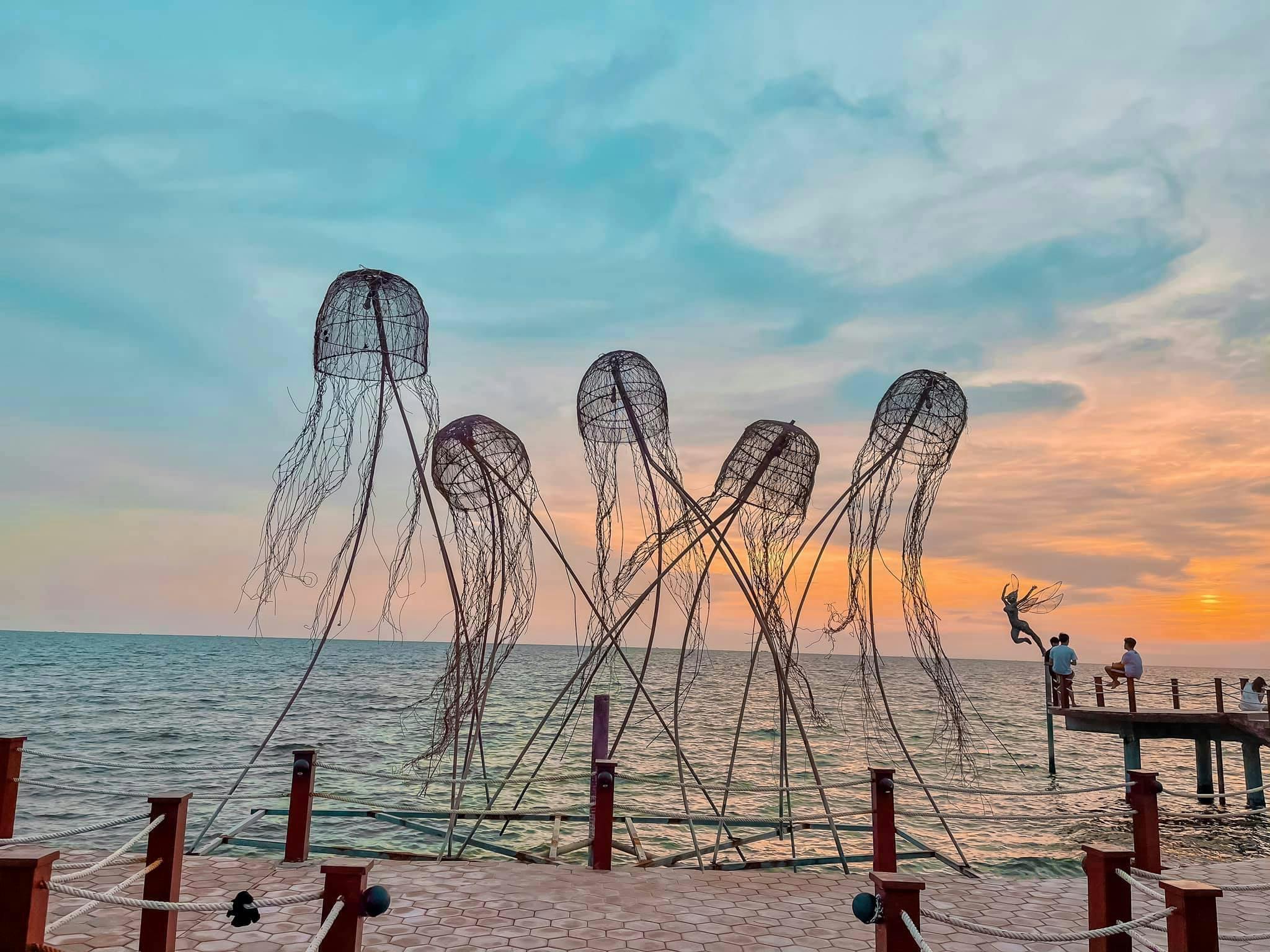 Artistic installations resembling jellyfish on a seaside pier at sunset, with a person standing in the distance.