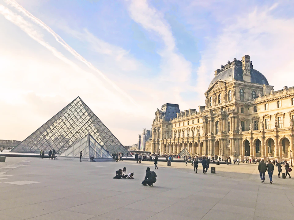 People are gathered around the glass pyramid entrance of the Louvre Museum in Paris, with the historic building in the background.