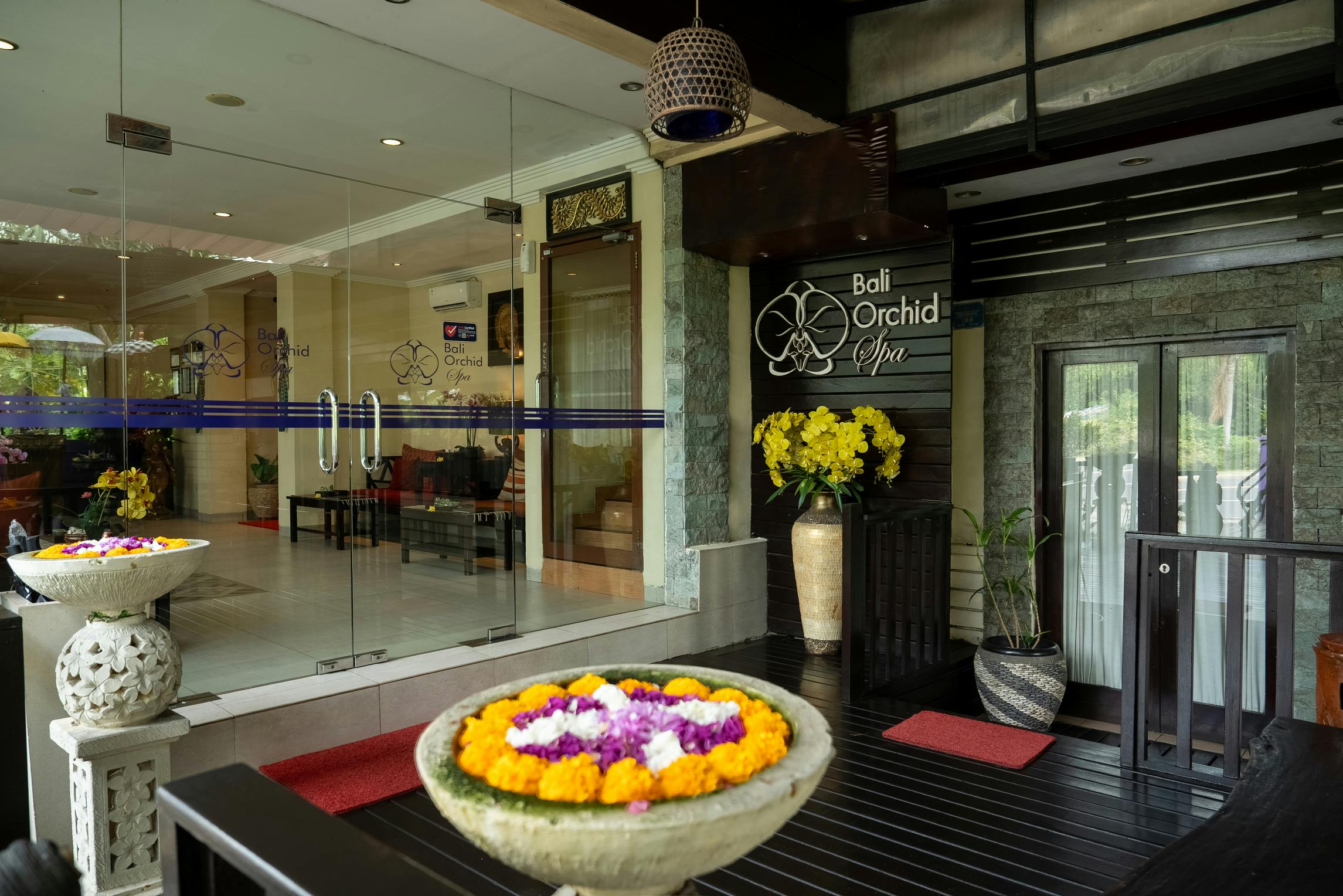 Wejście do Bali Orchid Spa z dekoracjami kwiatowymi, szklanymi drzwiami i roślinami w wazonach.