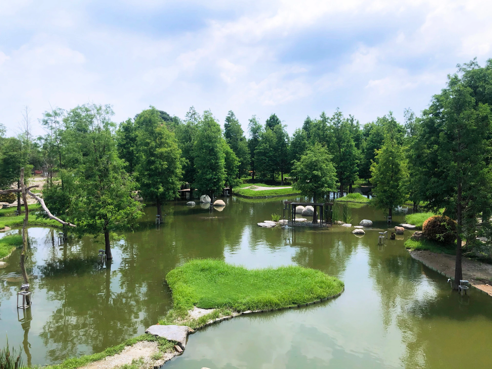 A tranquil park scene with trees, small grassy islands, and a pond under a partly cloudy sky.