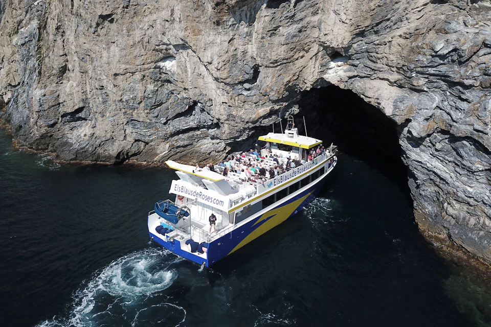 A tour boat carrying passengers approaches the entrance to a rocky cave on a calm stretch of water.