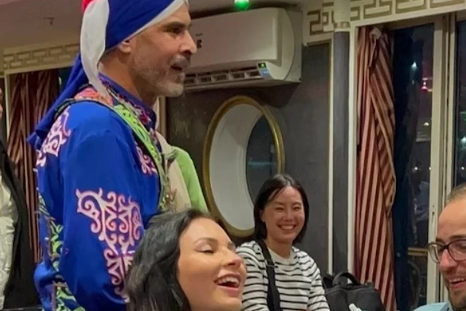 A man in colorful traditional attire interacts with seated people indoors, creating a lively and cheerful atmosphere.