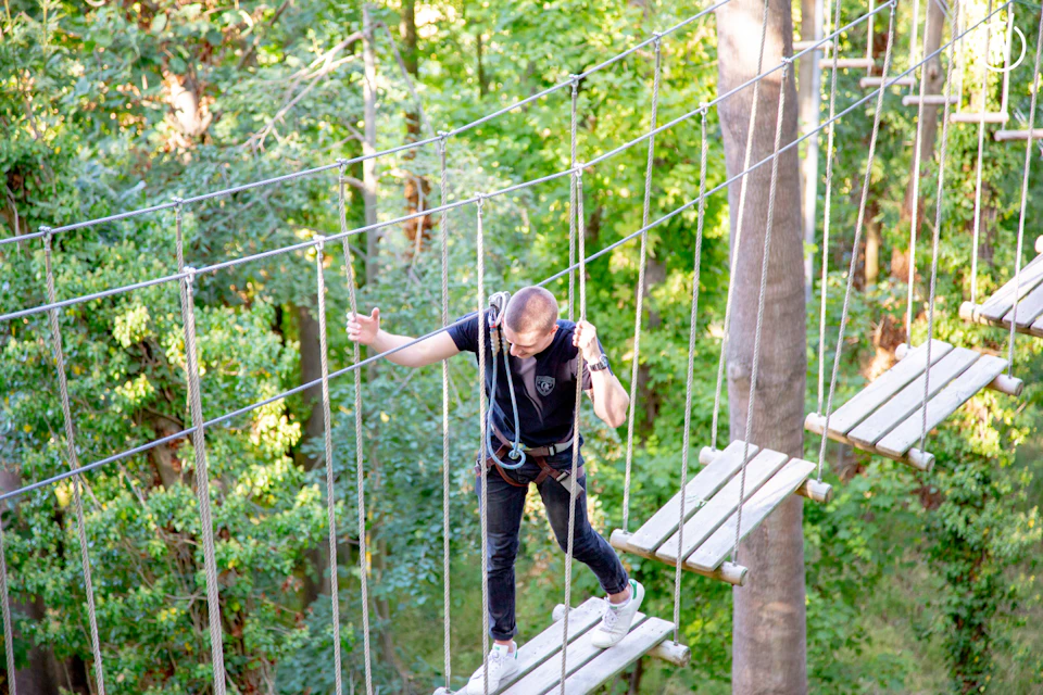 Person on a rope bridge holding onto ropes, surrounded by a green forest. They are wearing a harness and white shoes.