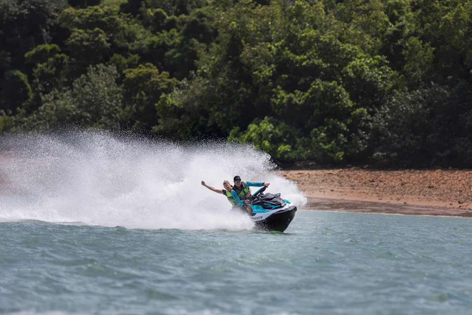 Two people riding a jet ski on water, making a splash with a tree-lined shoreline in the background.