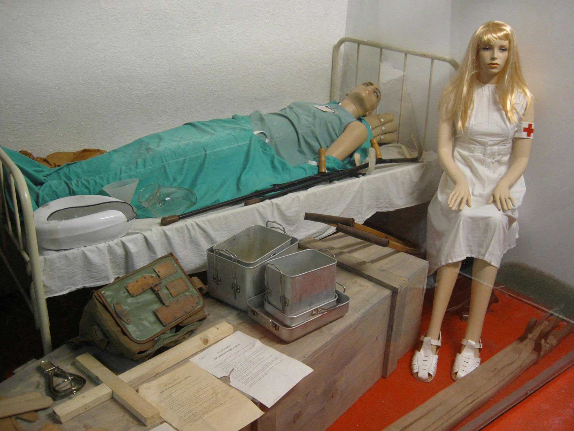 Scéna zobrazující vintage lékařské figuríny: jedna oblečená jako zdravotní sestra sedící na bedně a druhá na nemocničním lůžku. Zdravotnické potřeby v okolí.