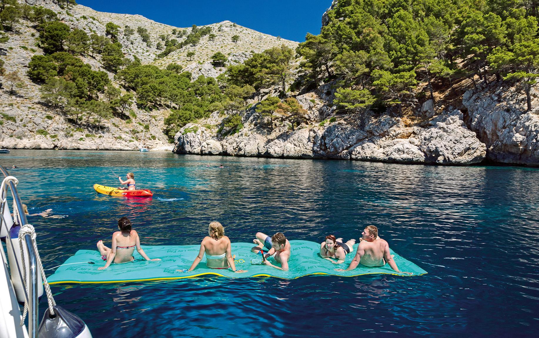 Cinq personnes se détendent sur un grand tapis flottant dans une eau bleue claire, avec des falaises rocheuses et une végétation luxuriante en arrière-plan.