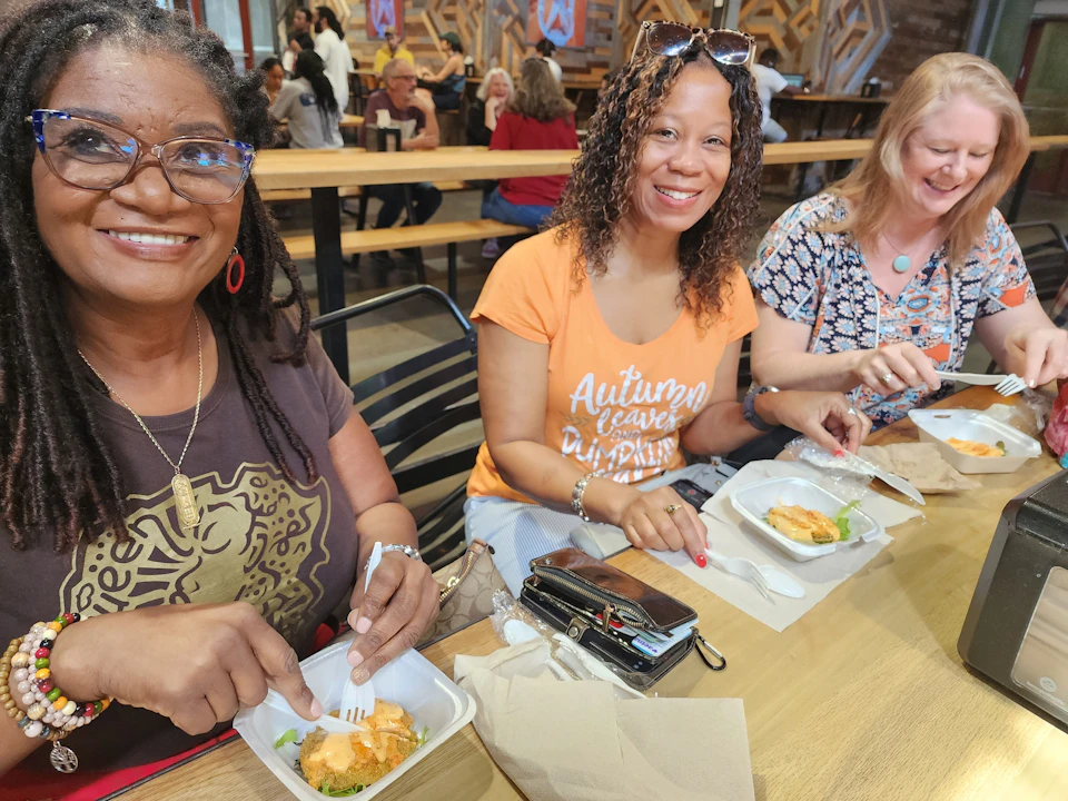 Two women smiling at a table in a casual dining setting, with plates of food and utensils in front of them.