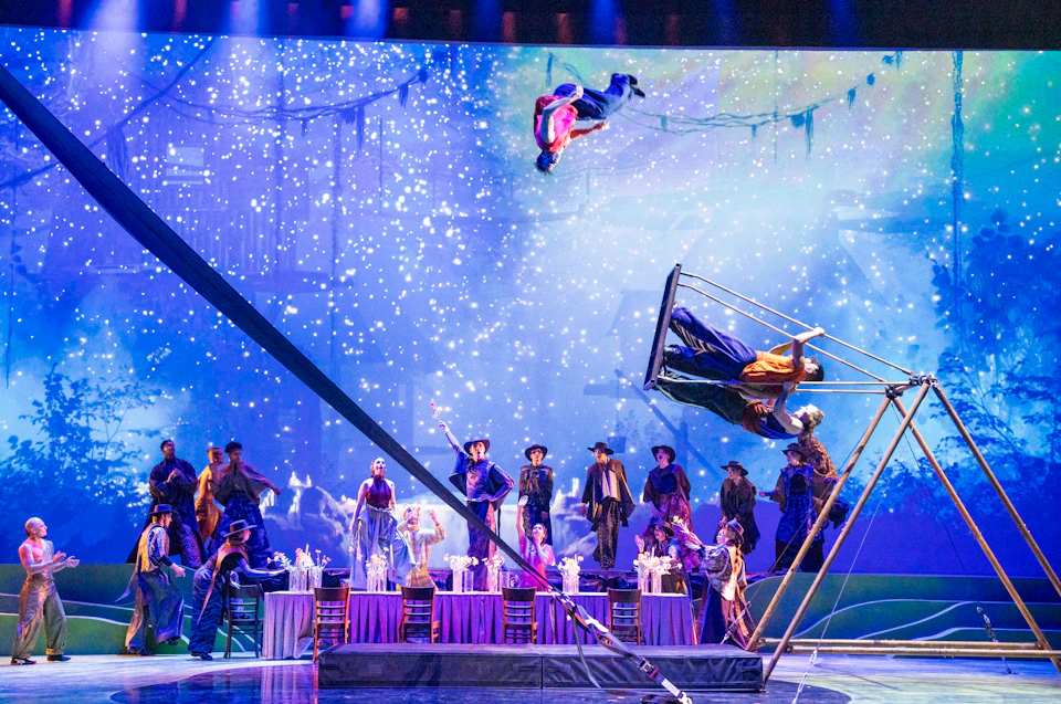 A vibrant performance with acrobats, a dinner table setup, and a starry background on a theatrical stage.