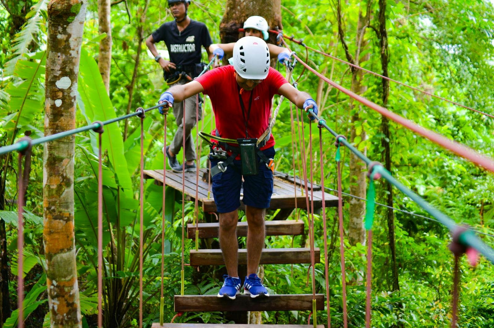 Dos personas navegando por un puente de cuerda en un frondoso bosque, con un instructor observando desde atrás. Ambos llevan cascos y arneses.