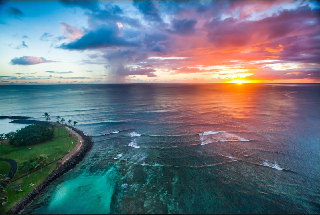 Aerial view of an ocean at sunset with vibrant orange and purple skies, calm waves, a coastline with palm trees, and a cloudburst.