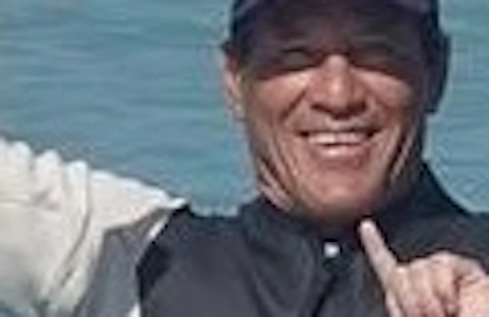 Three people sitting on surfboards in the water near a rocky shore, smiling and making hand gestures towards the camera.