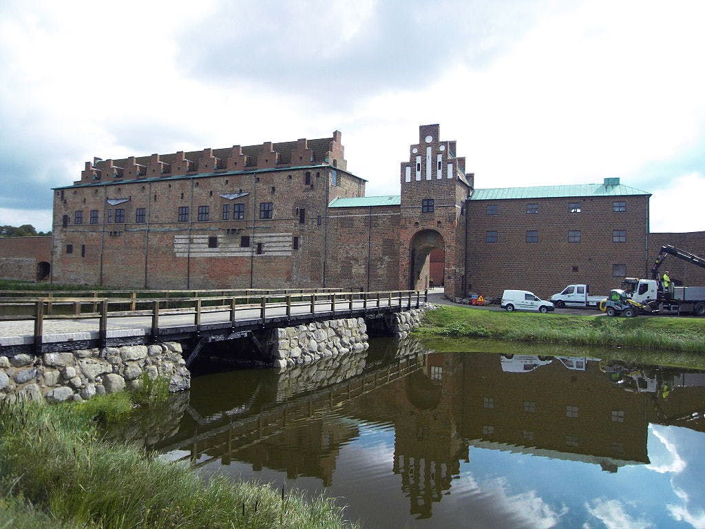 En historisk murstensbygning med et porttårn, der støder op til en stenbro over en voldgrav, med parkerede lastbiler og en kran i nærheden.
