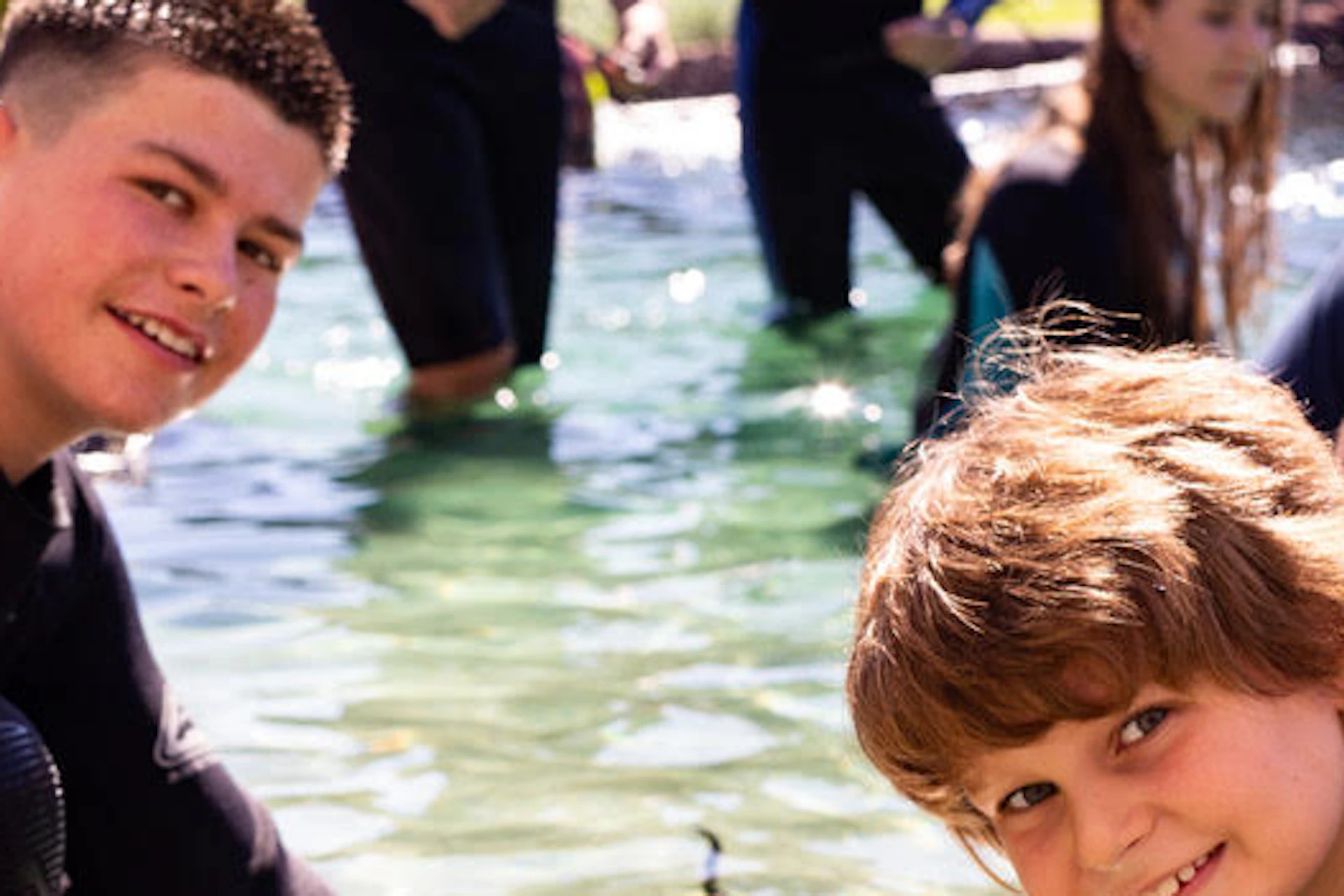 Two smiling boys pet a stingray in a shallow pool, with other people in the background participating in the activity.