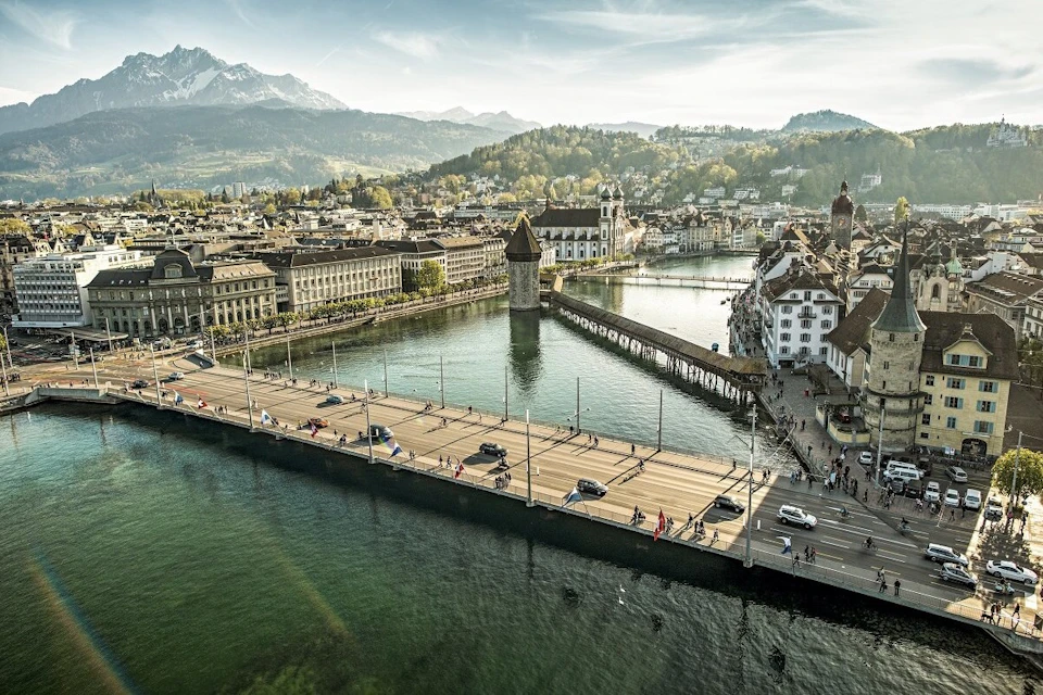 Aerial view of a city with a river, a bridge, a medieval tower, and picturesque buildings surrounded by mountains.