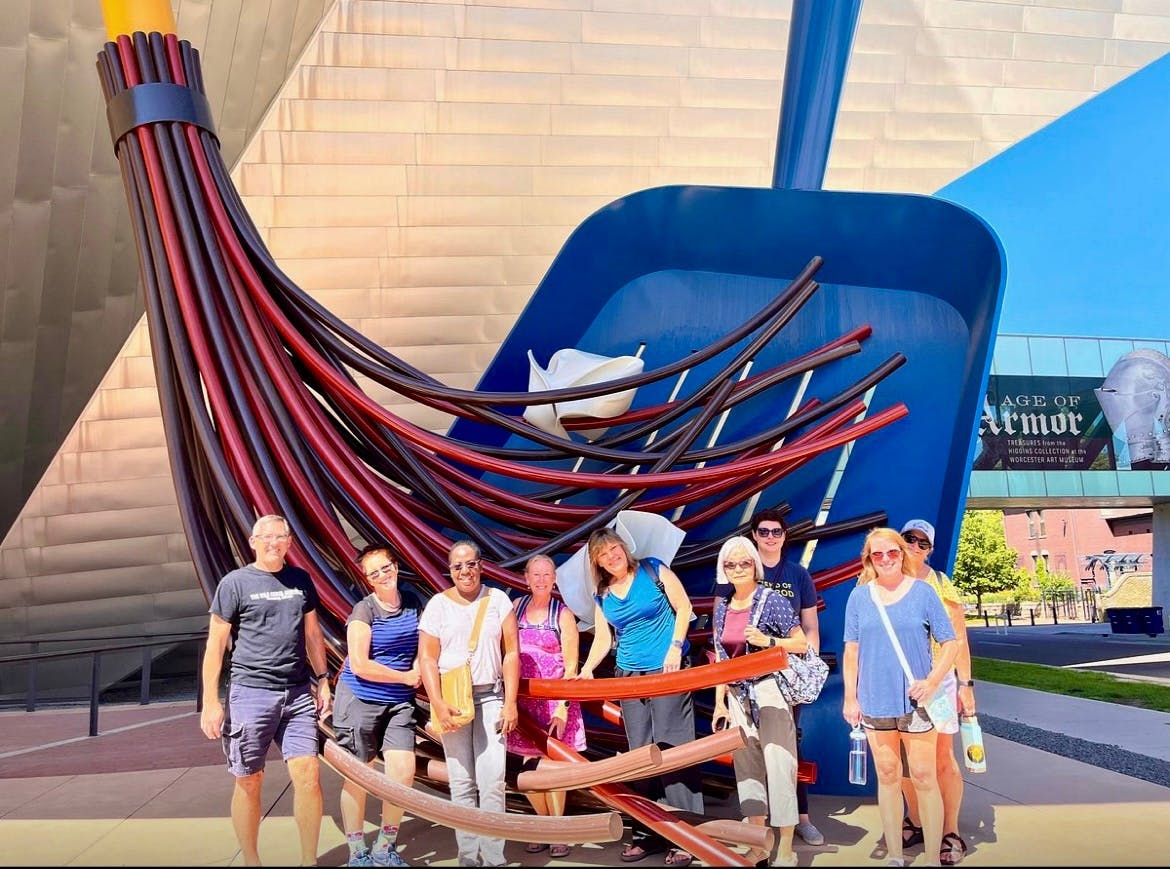 A group of people posing in front of a large sculpture resembling a stylized broom and dustpan outdoors.