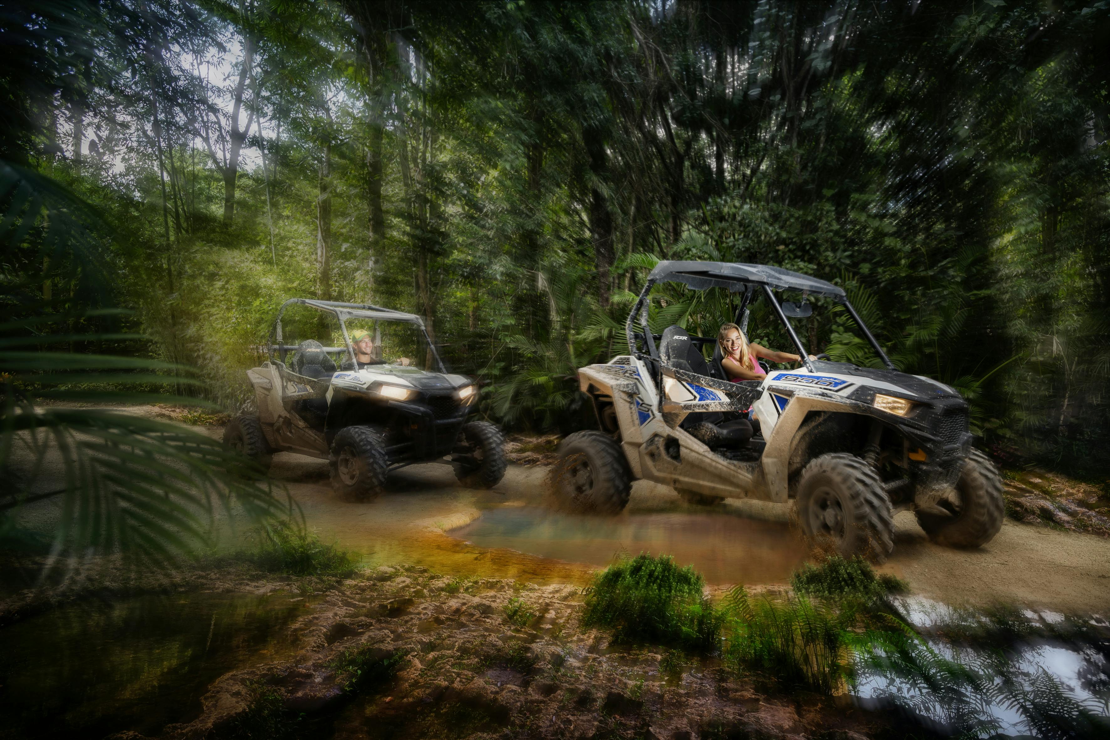 Δύο οχήματα εκτός δρόμου οδηγούν μέσα από ένα λασπωμένο δασικό μονοπάτι με καταπράσινα δέντρα. Κάθε όχημα έχει δύο άτομα μέσα.