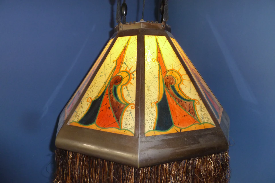 A stained glass lamp with geometric patterns in orange, yellow, and green, hangs from the ceiling against a blue wall.