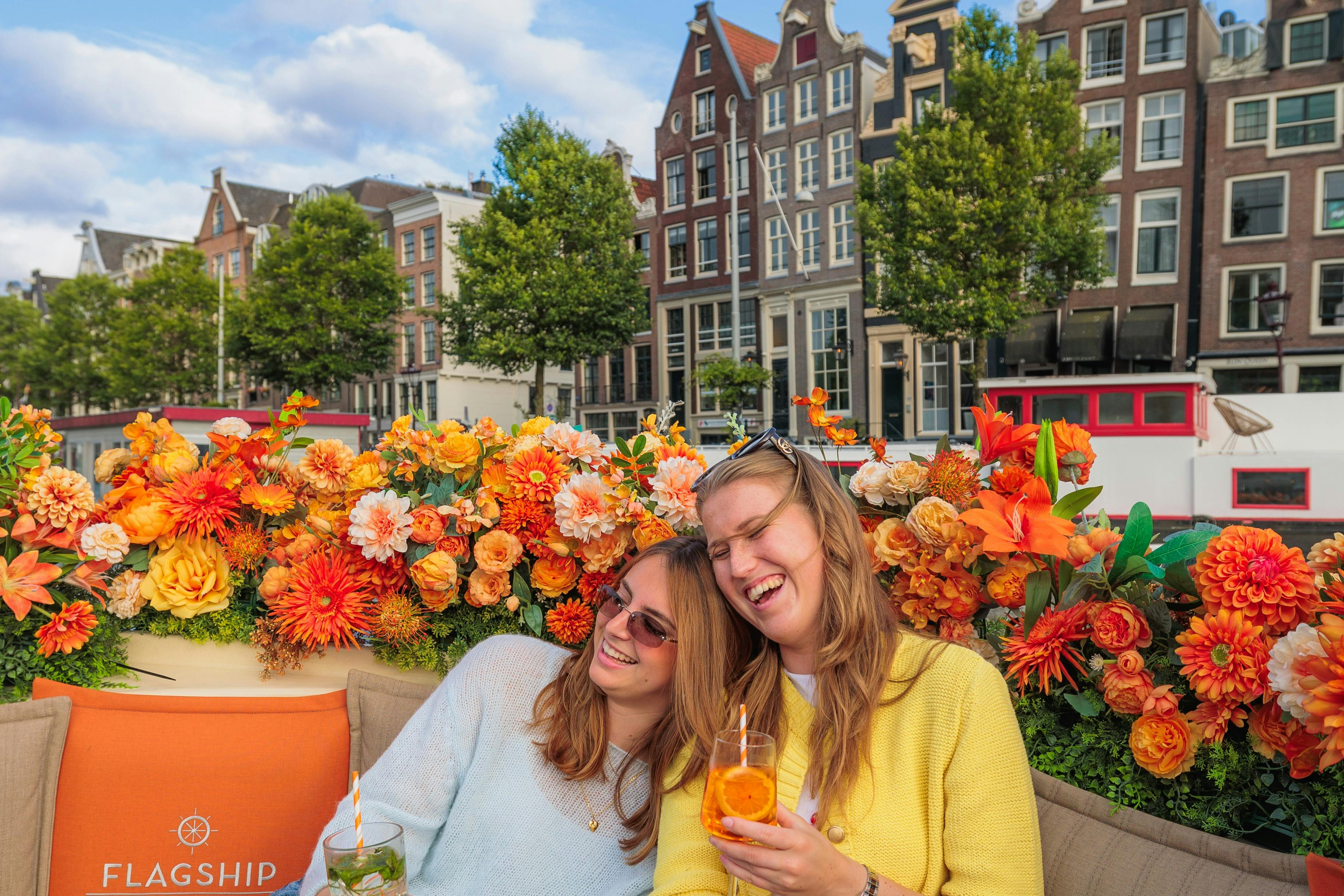 Two smiling women sit on a floral-decorated boat with orange flowers, enjoying drinks, with Dutch canal houses in the background.