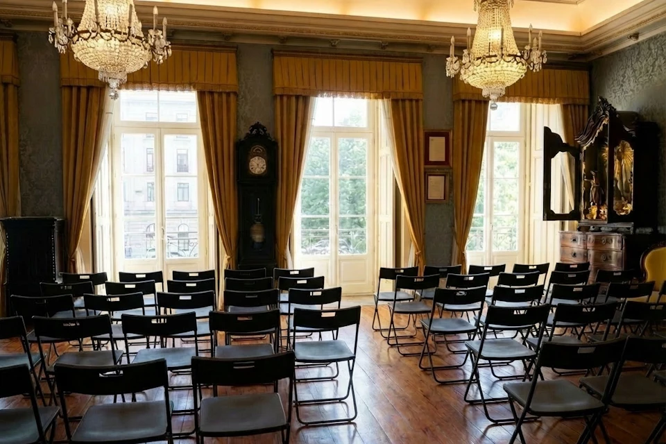 A room with rows of black chairs facing a set of large windows with yellow curtains, chandeliers, and a tall clock.