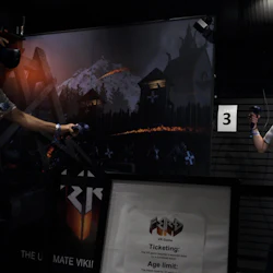 Two people wearing VR headsets and holding controllers in a dark room with Viking-themed decorations and a sign for a VR game.