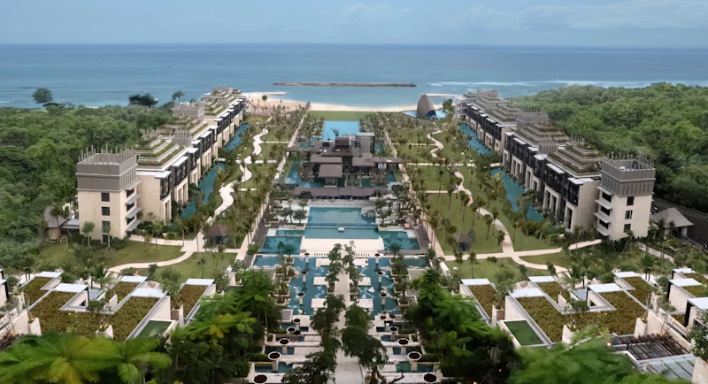 Aerial view of a resort with pools, gardens, and symmetrical buildings on both sides, facing a beach and the ocean.