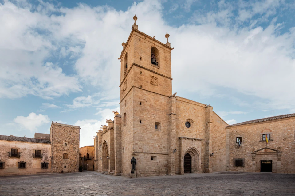 Historic stone church with a bell tower under a partly cloudy sky, surrounded by cobblestones and nearby buildings.