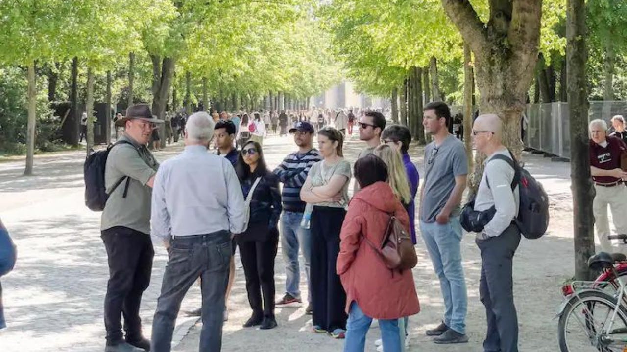 Skupina lidí stojí na stezce lemované stromy v parku a někteří z nich mají na sobě batohy a kola.