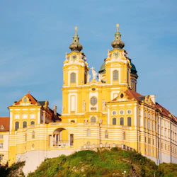 Gul byggnad i barockstil med två utsmyckade torn och detaljerad fasad, belägen på en grön kulle under en blå himmel.