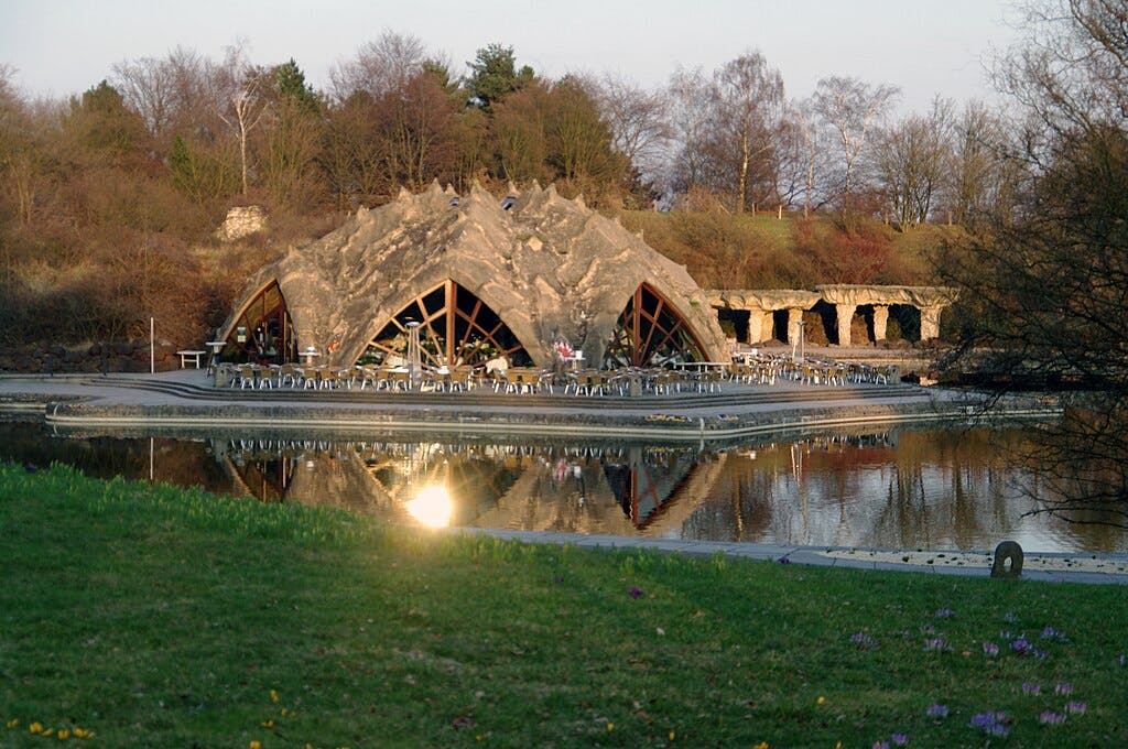 Stone building with large arches reflecting on the water, surrounded by trees and outdoor seating. Grassy foreground with flowers.