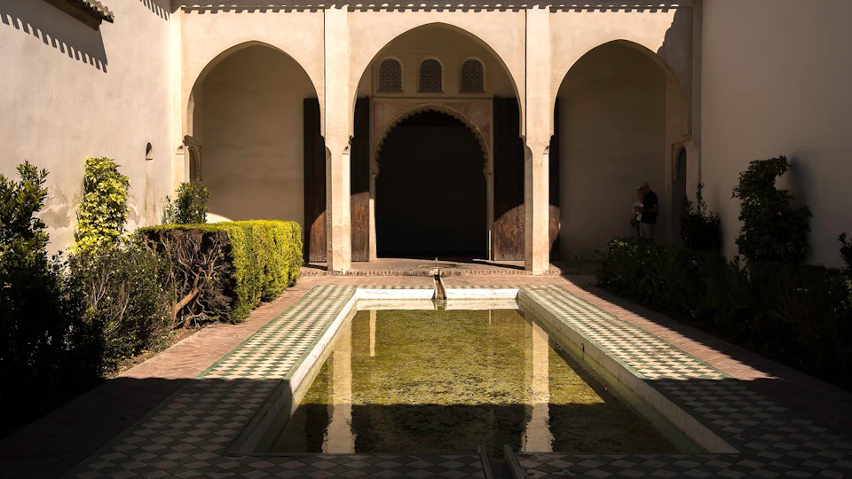 A courtyard with a rectangular reflecting pool, tiled floor, arched doorways, and trimmed greenery along the walls.