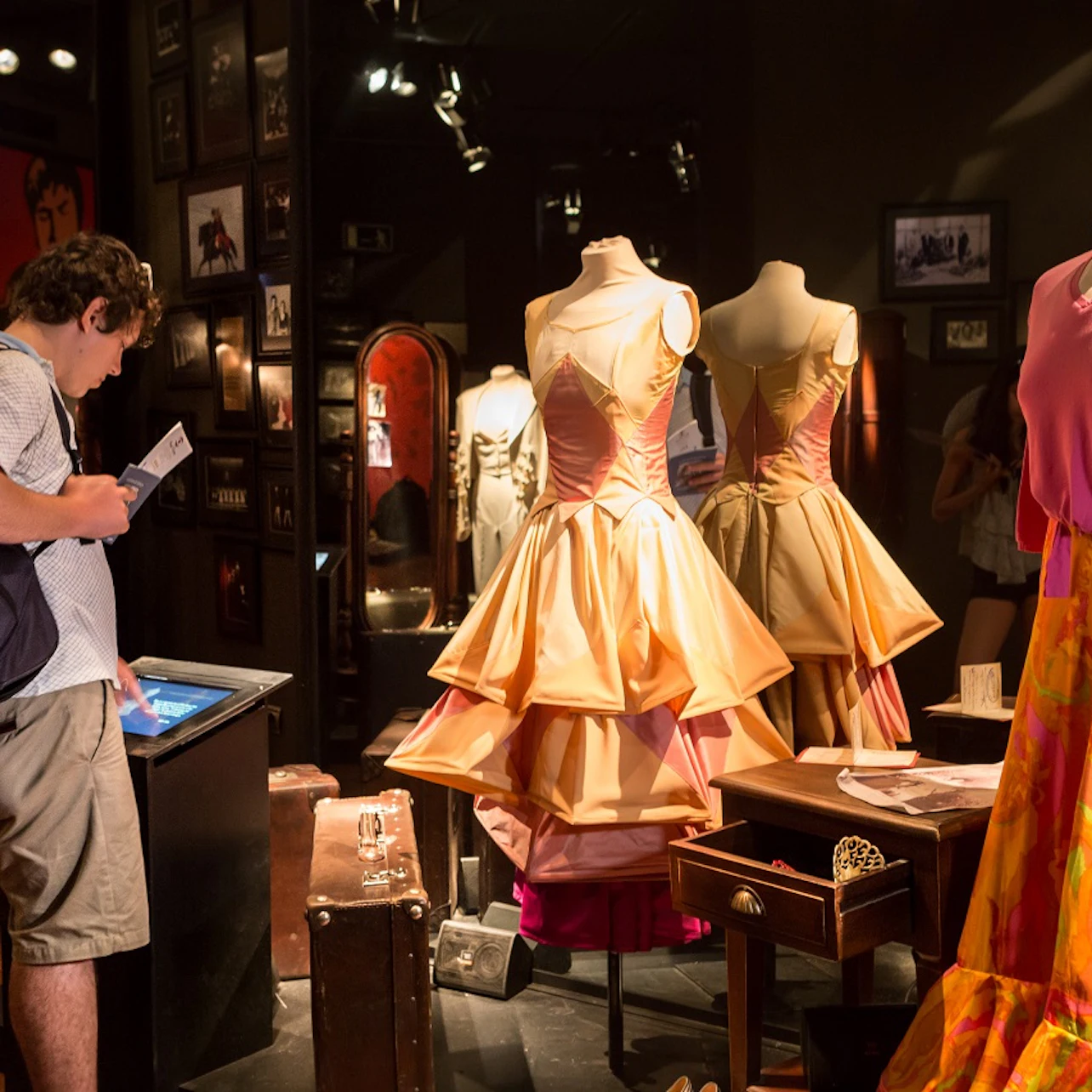 A person views an exhibit with mannequins displaying elaborate, layered dresses. Surrounding walls feature framed pictures and mirrors.